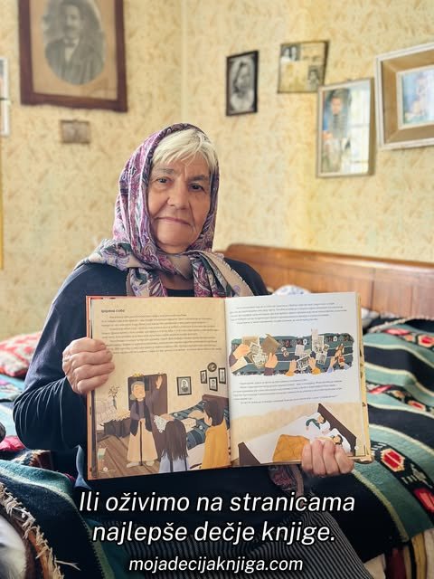 Nasmejana baka u šarenoj marami sedi na krevetu i drži otvorenu ilustrovanu dečju knjigu.