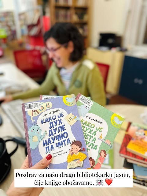 Ruka drži dve knjige Jasne Brkić, a u zamućenoj pozadini se smeje bibliotekarka u zelenom džemperu.