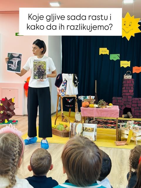 Nevena pokazuje dve fotografije gljiva i objašnjava deci kako da ih razlikuju, dok mališani pažljivo slušaju.
