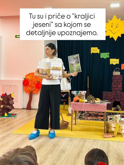 Dok drži otvorenu knjigu i veliku ilustraciju, Nevena deci priča o kraljici jeseni u vrtićkoj sali.