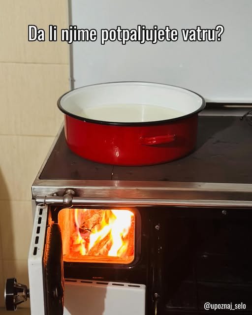 Crveni emajlirani lonac stoji na šporetu na drva, a kroz otvorena vratanca vidi se vatra.