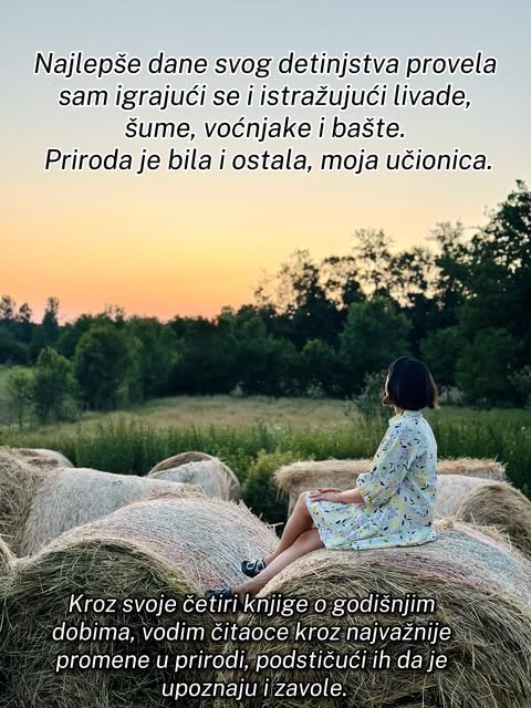 Žena u cvetnoj haljini posmatra livadu i šumu sa bale sena dok nebo narandžasto zasvetli pri zalasku.
