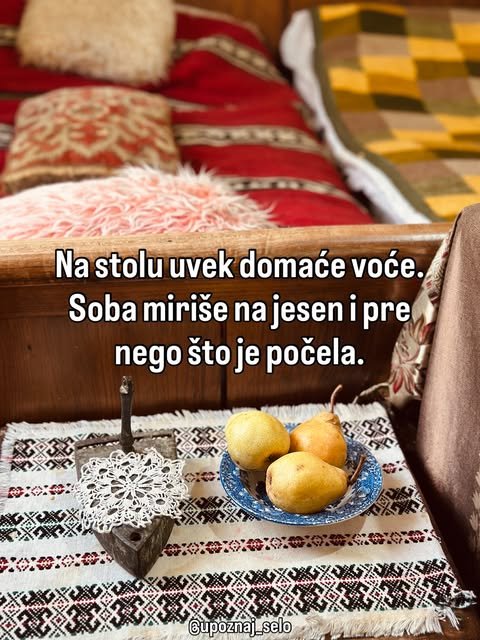 Tanjir sa tri kruške na tkanom stolnjaku pored stare gvozdene pegle, iza njih je krevet sa crvenim jastucima.