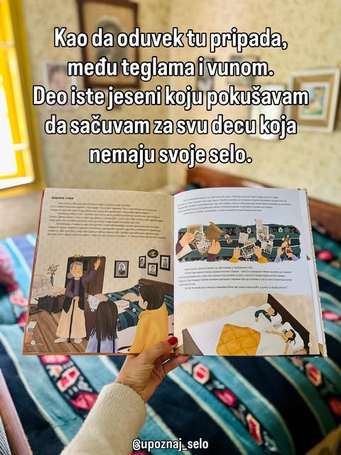 Ruka drži otvorenu ilustrovanu knjigu u sobi, ispod nje je krevet sa šarenim prekrivačem od vune.