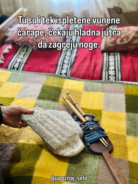 Ruka drži tek upletenu sivu čarapu, a pored nje na karirano ćebe leže drvena vretena sa vunom.