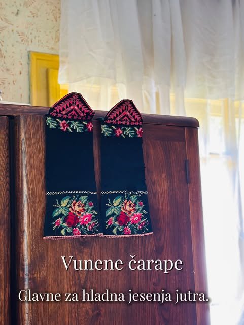 Dve crne vunene čarape sa vezenim ružama stoje uspravno na ormaru ispred prozora sa belom zavesom.