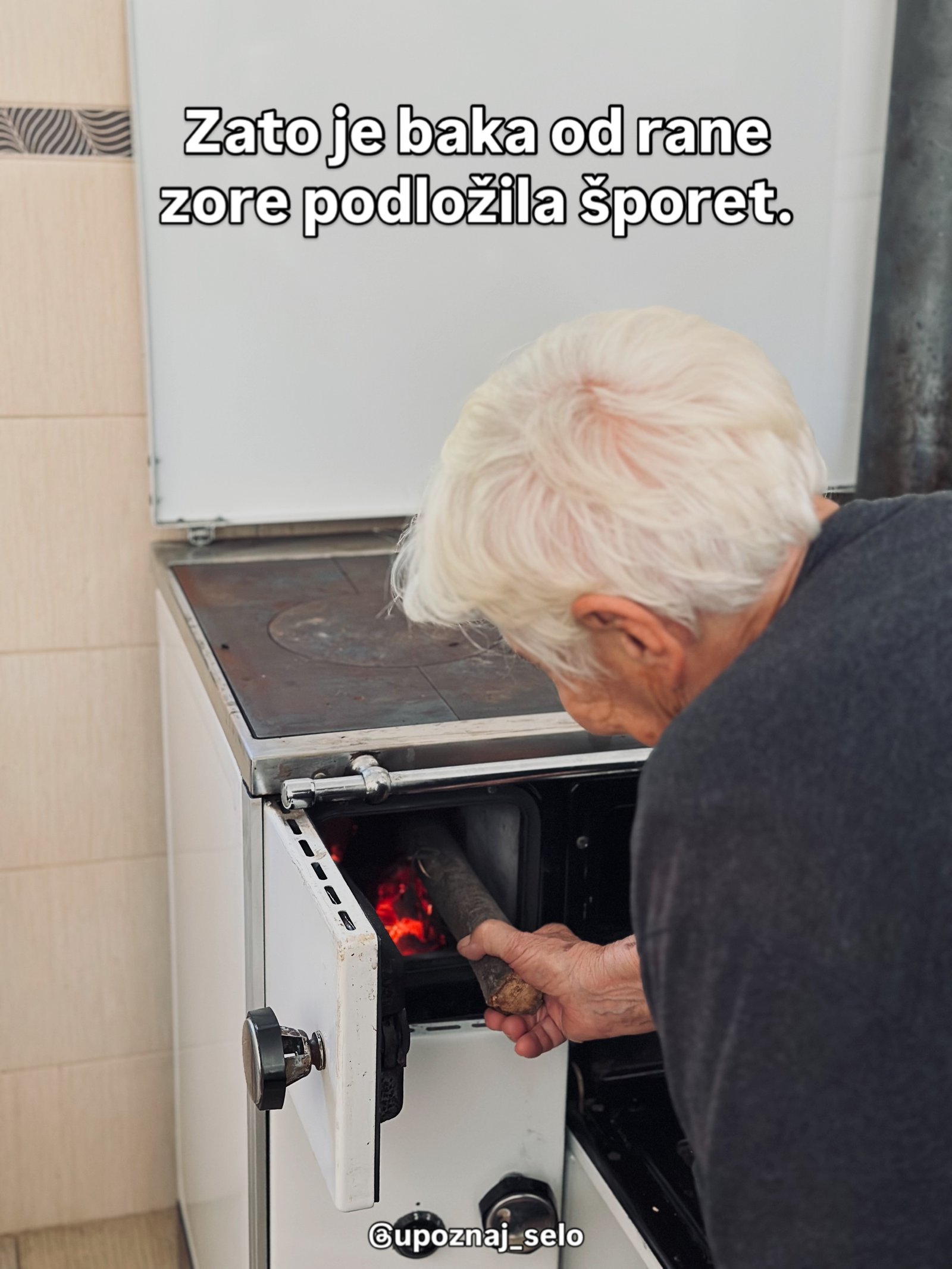 Sedokosa žena ubacuje cepanicu u ložište belog šporeta u kome gori vatra.