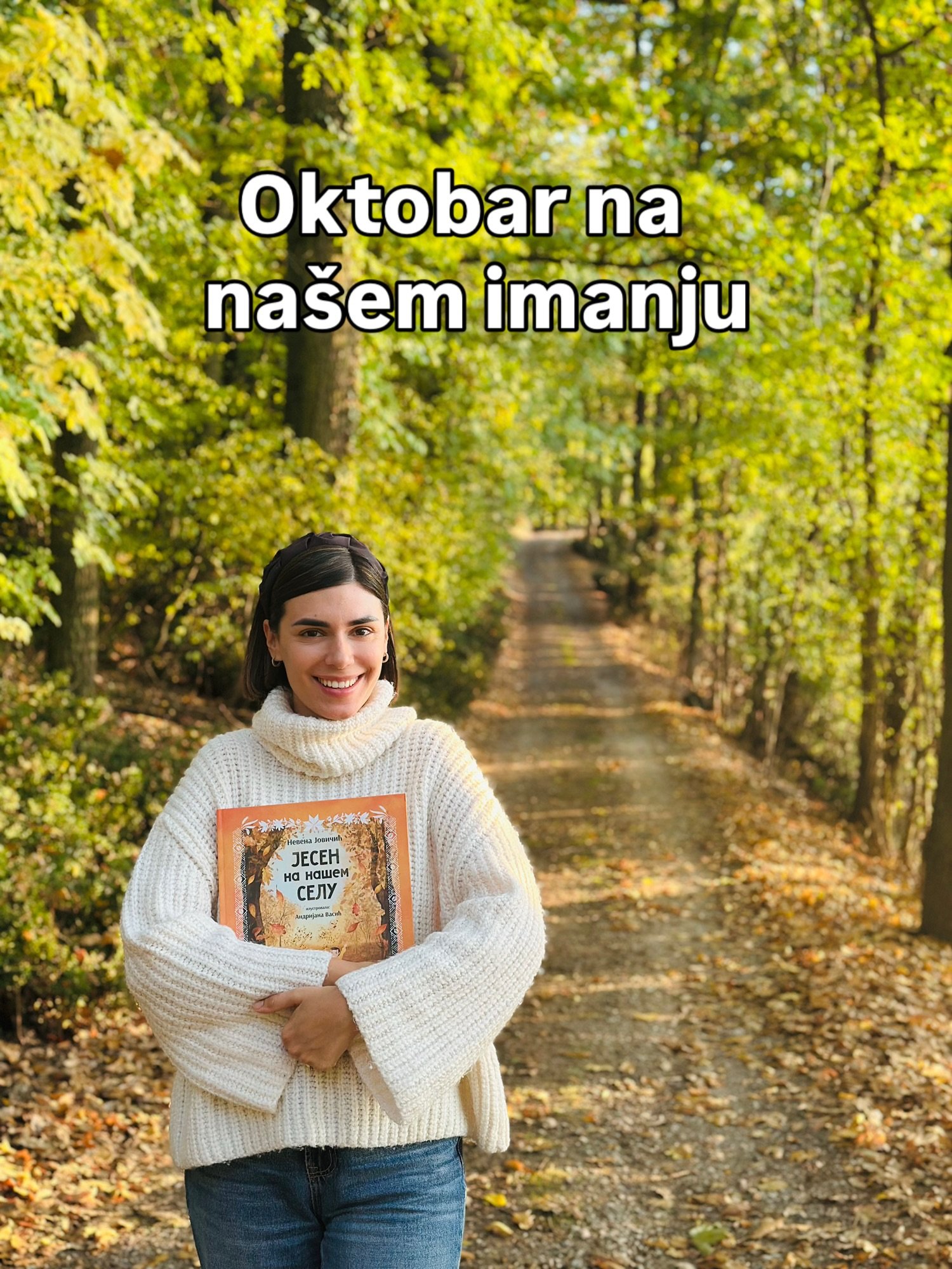 Nasmejana žena u belom pletenom džemperu drži knjigu „Jesen na našem selu“ na putu kroz šumu.