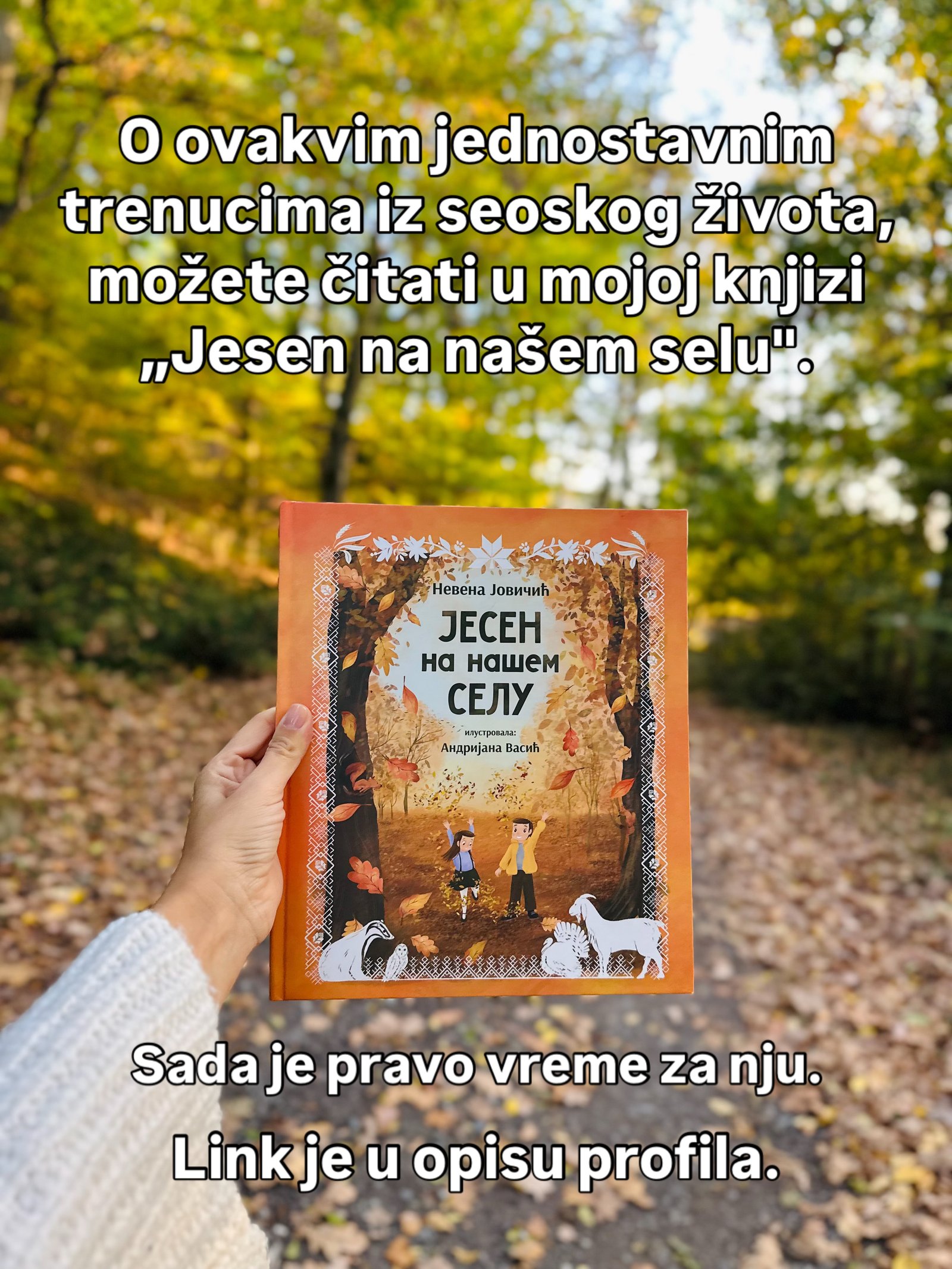 Ruka u belom pletenom rukavu drži knjigu narandžastih korica nad šumskim putem punim lišća.