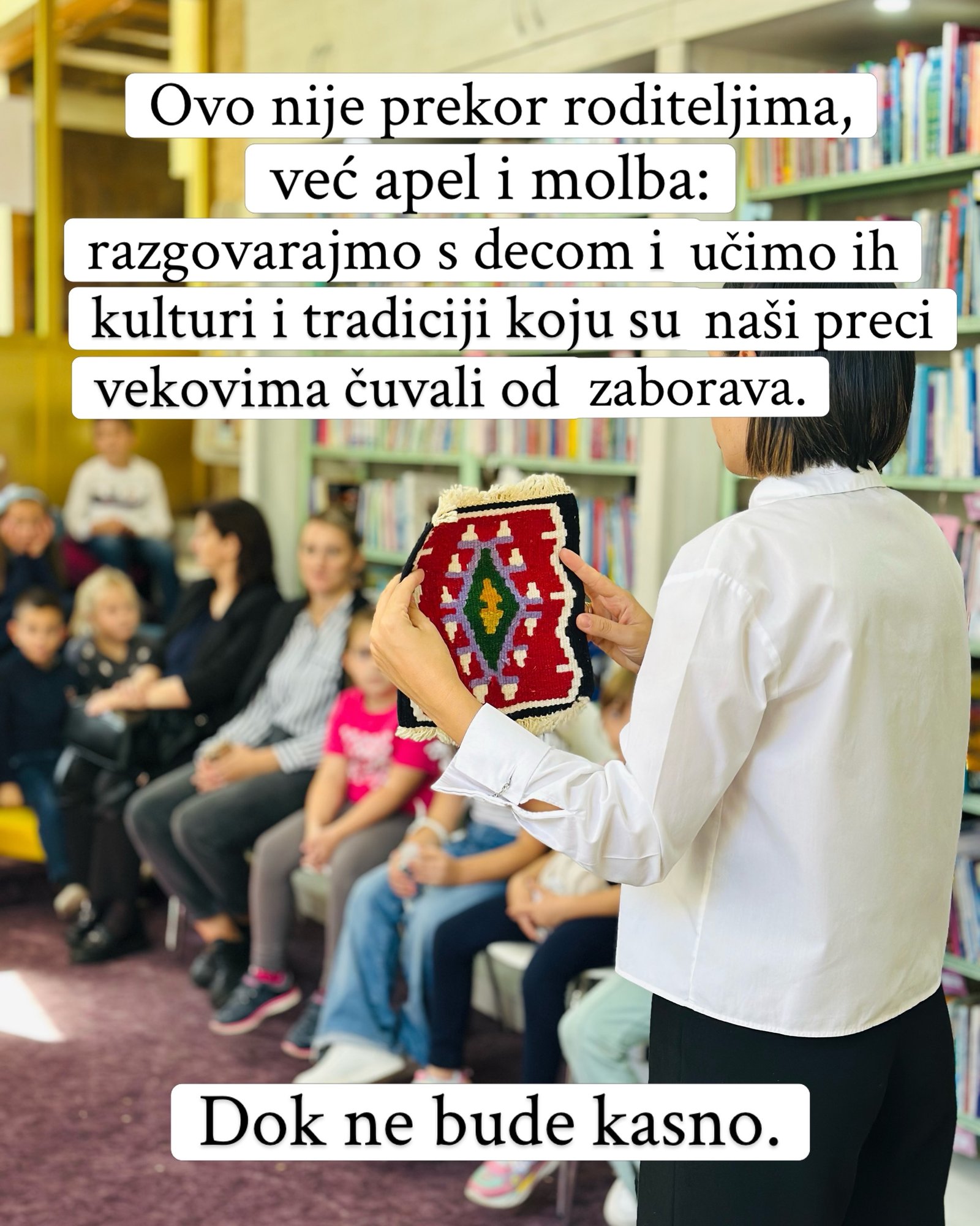 Žena u beloj košulji pokazuje mali tkani ćilim deci i roditeljima u biblioteci.