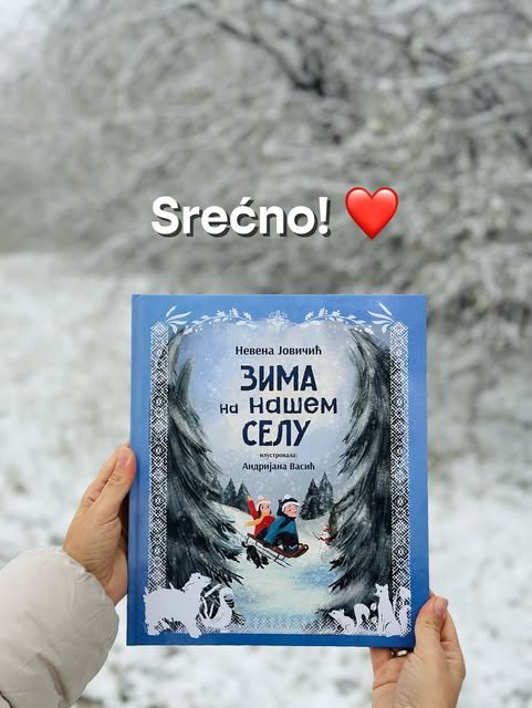 Ruke drže plavu slikovnicu „Zima na našem selu“ ispred drveća prekrivenog snegom i belog zimskog pejzaža.