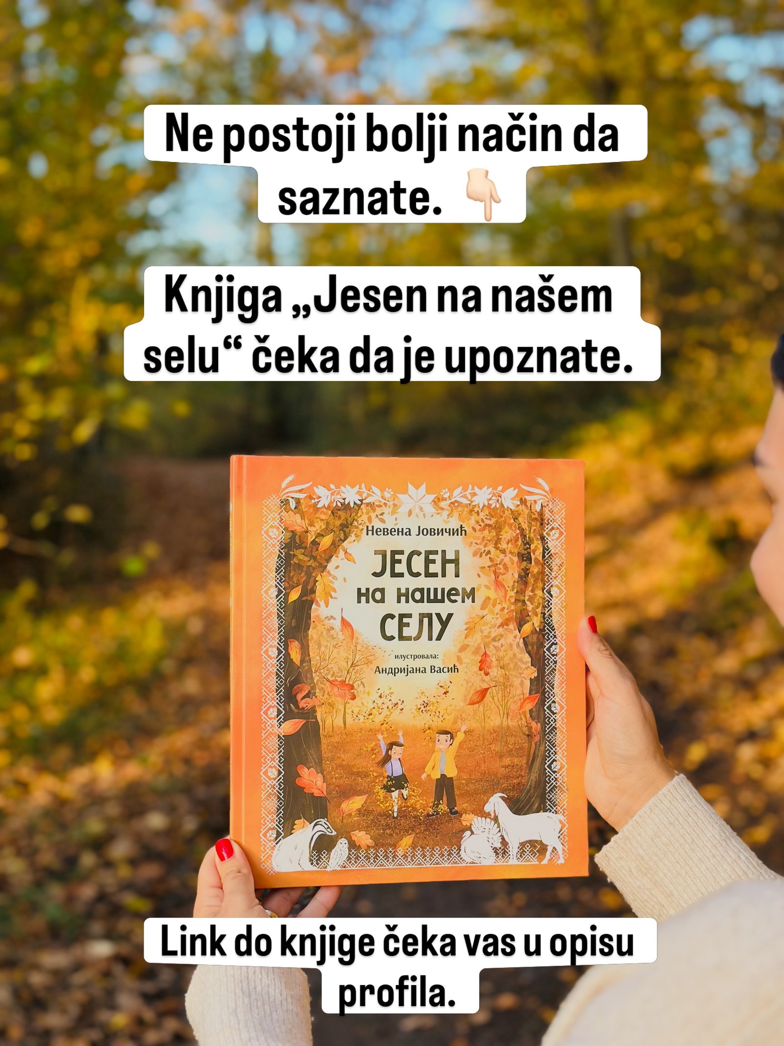 Ruke drže knjigu „Jesen na našem selu“ ispred šume u jesenjim bojama.