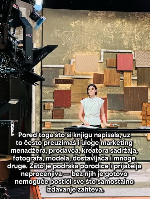 Autorka u beloj bluzi stoji za pultom u televizijskom studiju ispred zida od drvenih i crvenih panela.