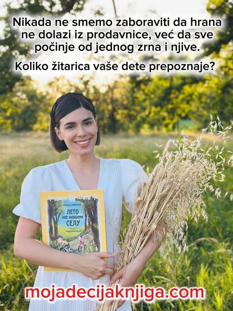 Nasmejana autorka u beloj haljini drži svoju dečju knjigu i snop ovsa na zelenoj livadi.
