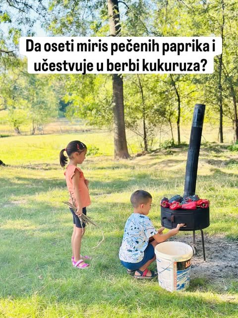Dečak i devojčica stoje na livadi pored crnog roštilja na kome se peku crvene paprike.